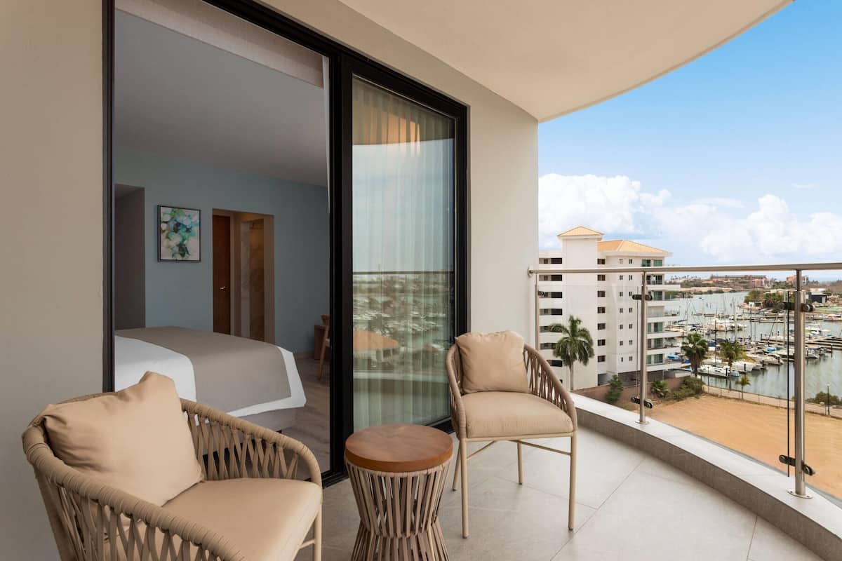 A balcony with wicker chairs, a small round table, and a bed with a white headboard. A large window offers a view of a marina with boats and buildings.