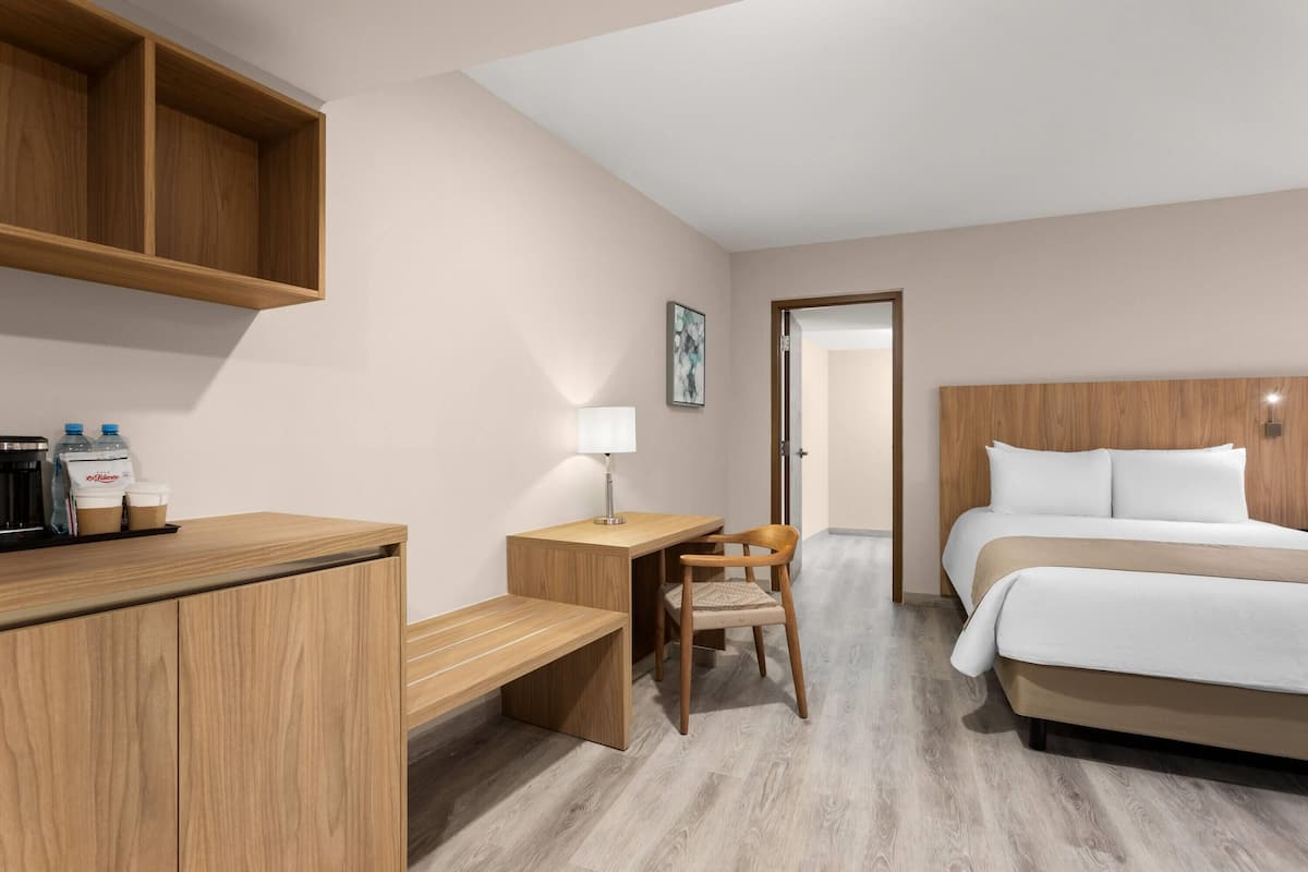 A hotel room with a bed, a desk with a chair, a wooden cabinet, and a door leading to another room.