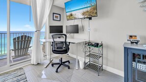 A room with a desk, chair, TV, and a balcony with a view of the beach. - Shores of Panama 1002 (Panama City Beach)