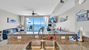 A modern kitchen with a granite countertop, double sink, and a view of the ocean through the sliding glass door. - Shores of Panama 1002 (Panama City Beach)