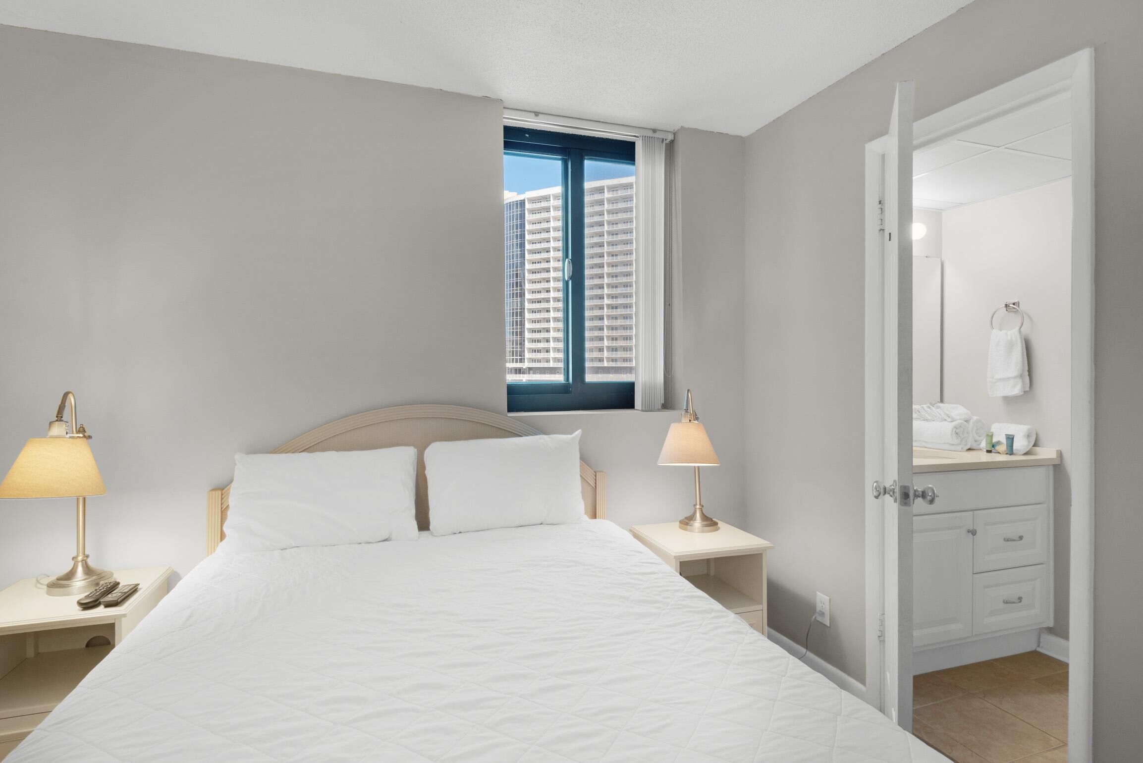 Habitación de hotel con cama, dos lámparas de noche, ventana con vistas a edificios altos y baño con espejo y toallero.