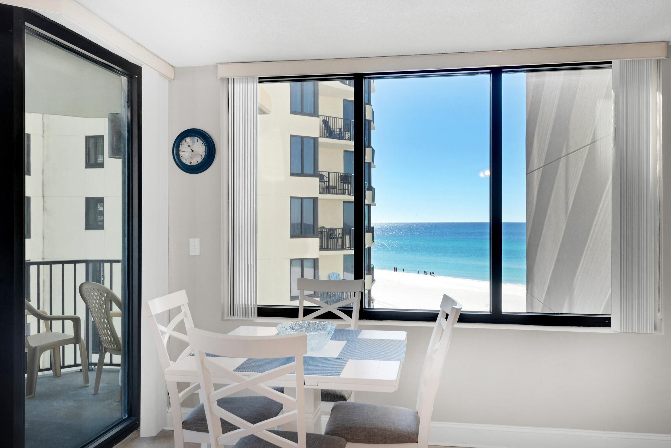 Ein Zimmer mit Blick auf den Strand, ein Tisch mit Stühlen, eine Uhr an der Wand und ein Balkon mit Sitzgelegenheiten.