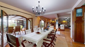 A dining area with a long table, chairs, and a chandelier. There is a kitchen in the background with stone walls and wooden cabinets. - Primavera Villa Sea View (Aigialeia)