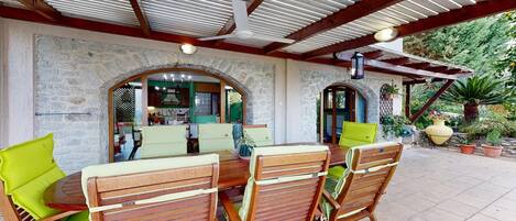 An outdoor patio with a stone wall, wooden furniture, and a ceiling fan.