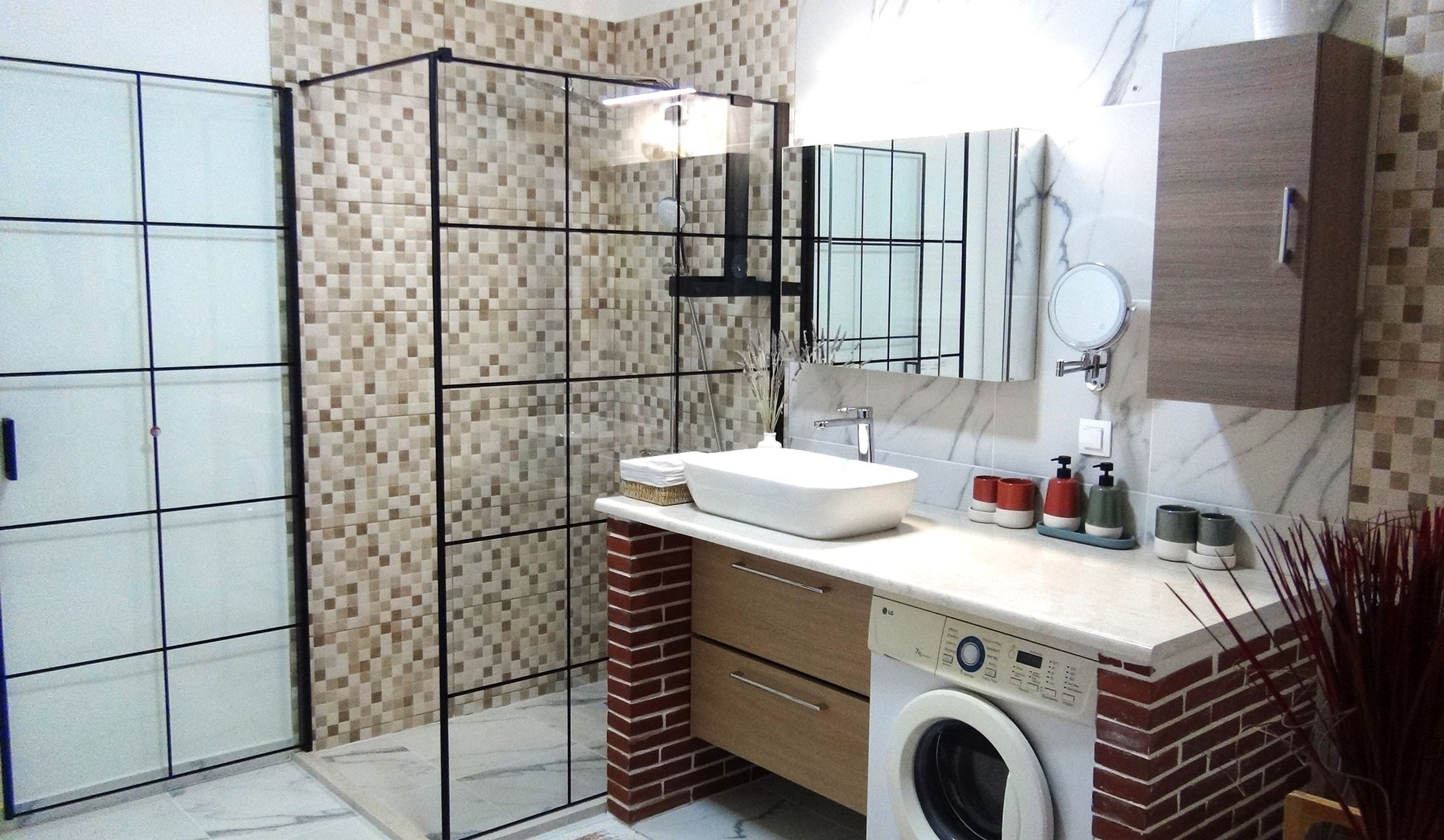 A bathroom with a tiled shower enclosure, a sink with a mirror, a washing machine, and a cabinet.