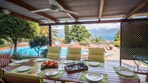 A covered outdoor dining area with a table set for four, overlooking a pool and scenic views. - Primavera Villa Sea View (Aigialeia)