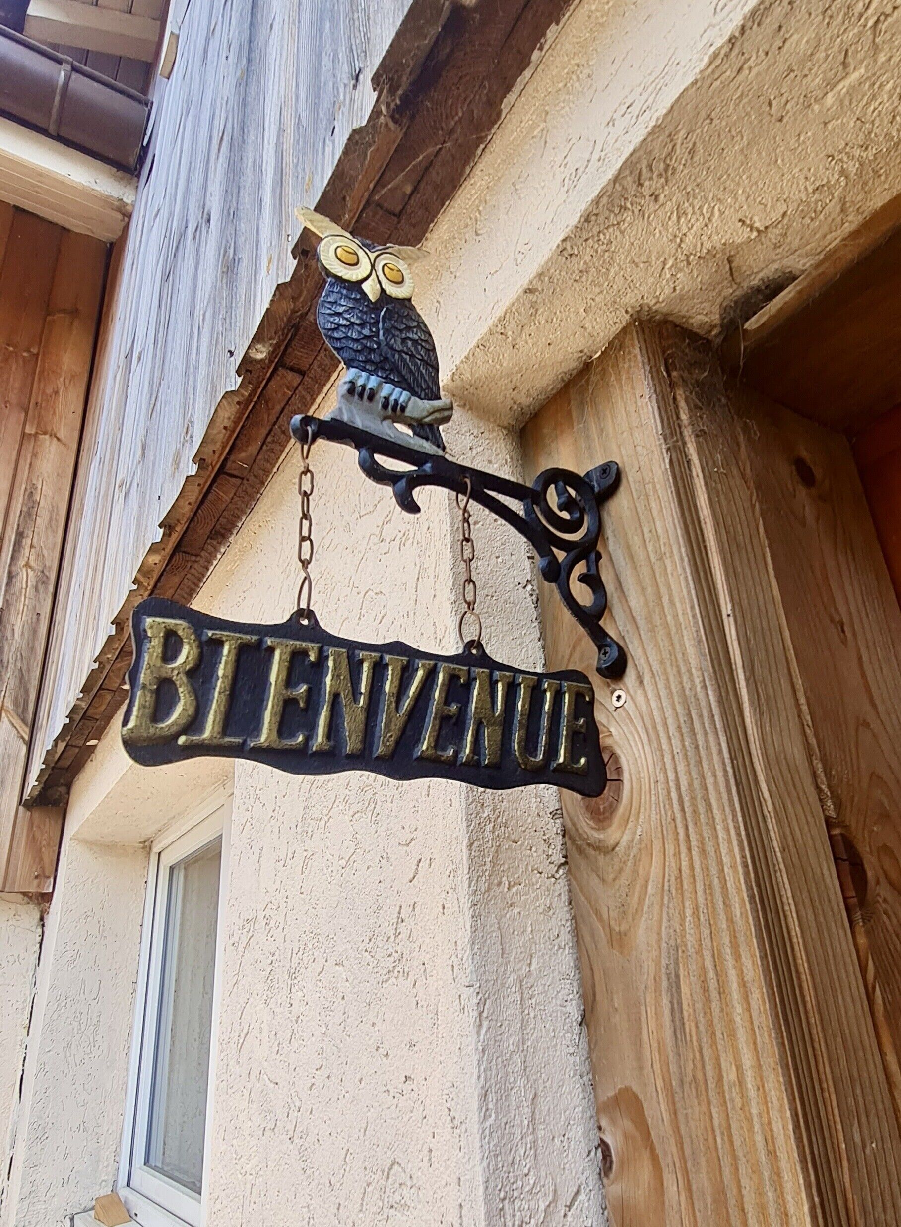 A wooden exterior with a 'BIENVENUE' sign hanging from a metal fixture, an owl decoration above it, and a window to the left.