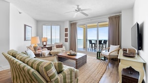 A spacious living room with a large sofa, a coffee table, and a TV on a stand. There is a balcony with a table and chairs outside.