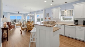 A modern kitchen with white cabinetry, a central island, and a dining area with wooden chairs and a table. - Riviera Dunes 203 (Pensacola)
