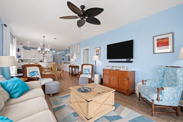 Ein Wohnzimmer mit einer Couch, Hocker, Couchtisch aus Holz, Korbstühlen, einem Fernseher und einem Deckenventilator.