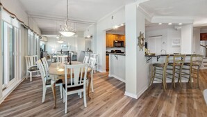 Un grand espace dédié à la salle à manger, avec une table ronde et des chaises blanches, une cuisine dotée de meubles en bois et un coin bar avec des tabourets.