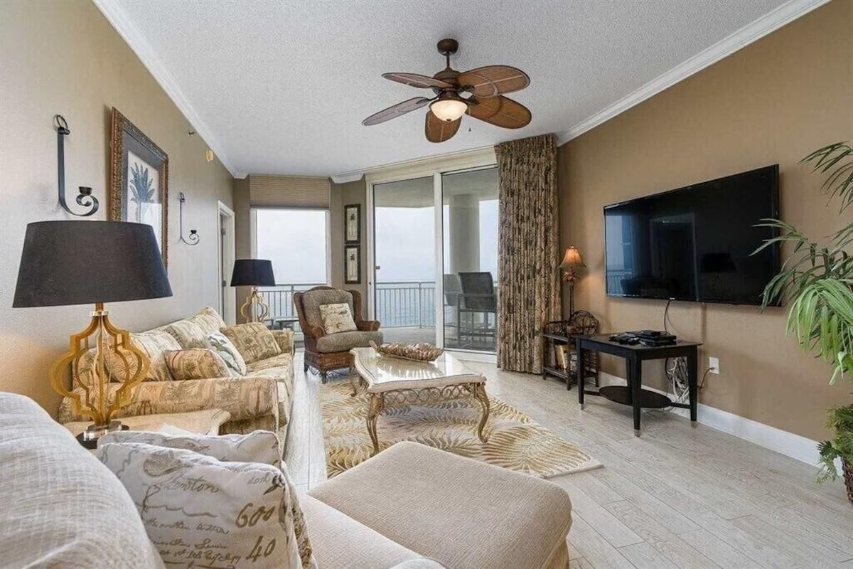 A spacious living room with a ceiling fan, a flat-screen TV, and a large window offering a view of the outdoors.