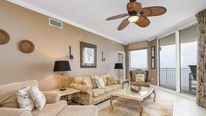A spacious living room with a beige sofa, a matching armchair, a coffee table, and a ceiling fan. There is a large window with a view of the outdoors. - Palacio 1202 (Pensacola)