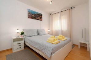 Un dormitorio ordenado con cama, dos mesitas de noche, una lámpara, una planta pequeña, un cuadro y un televisor.
