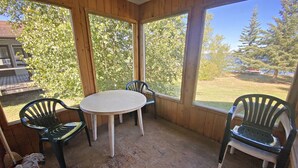 Eine überdachte Terrasse mit rundem Tisch und vier Stühlen, großen Fenstern und Blick auf Bäume und ein Gebäude.