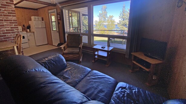Ein gemütliches Wohnzimmer mit einem blauen Ledersofa, einem kleinen Tisch und einem Fernseher. Es gibt eine Küchenzeile mit einem Kühlschrank und einer Ziegelwand. Das Zimmer verfügt über große Fenster mit Blick auf Bäume und eine Terrasse.