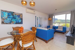 A modern living room with a glass dining table, wicker chairs, a blue sofa, and a large abstract painting on the wall.