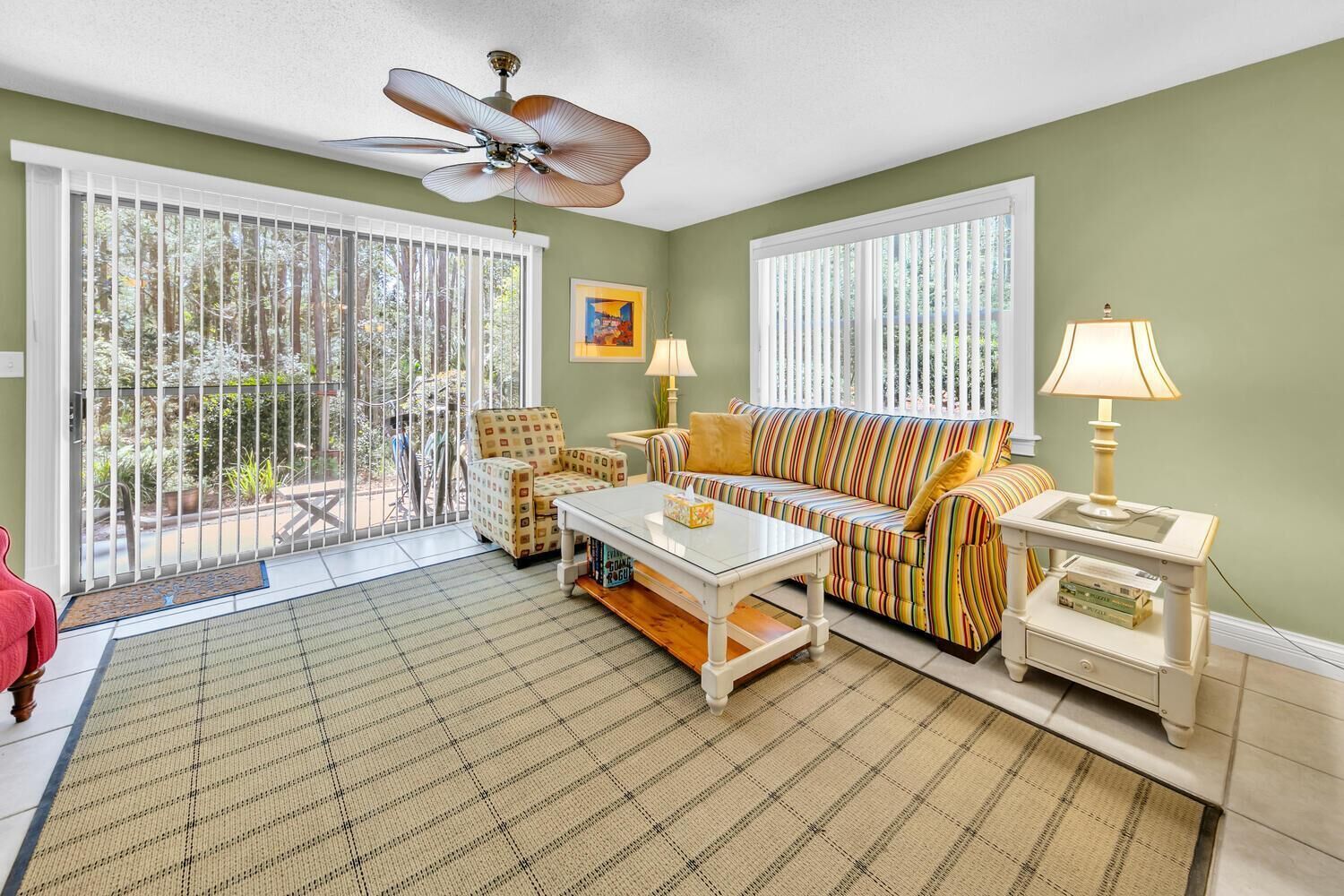 A living room with a sofa, armchairs, a coffee table, and a lamp. There is a ceiling fan, a window with blinds, and a door leading to a balcony.