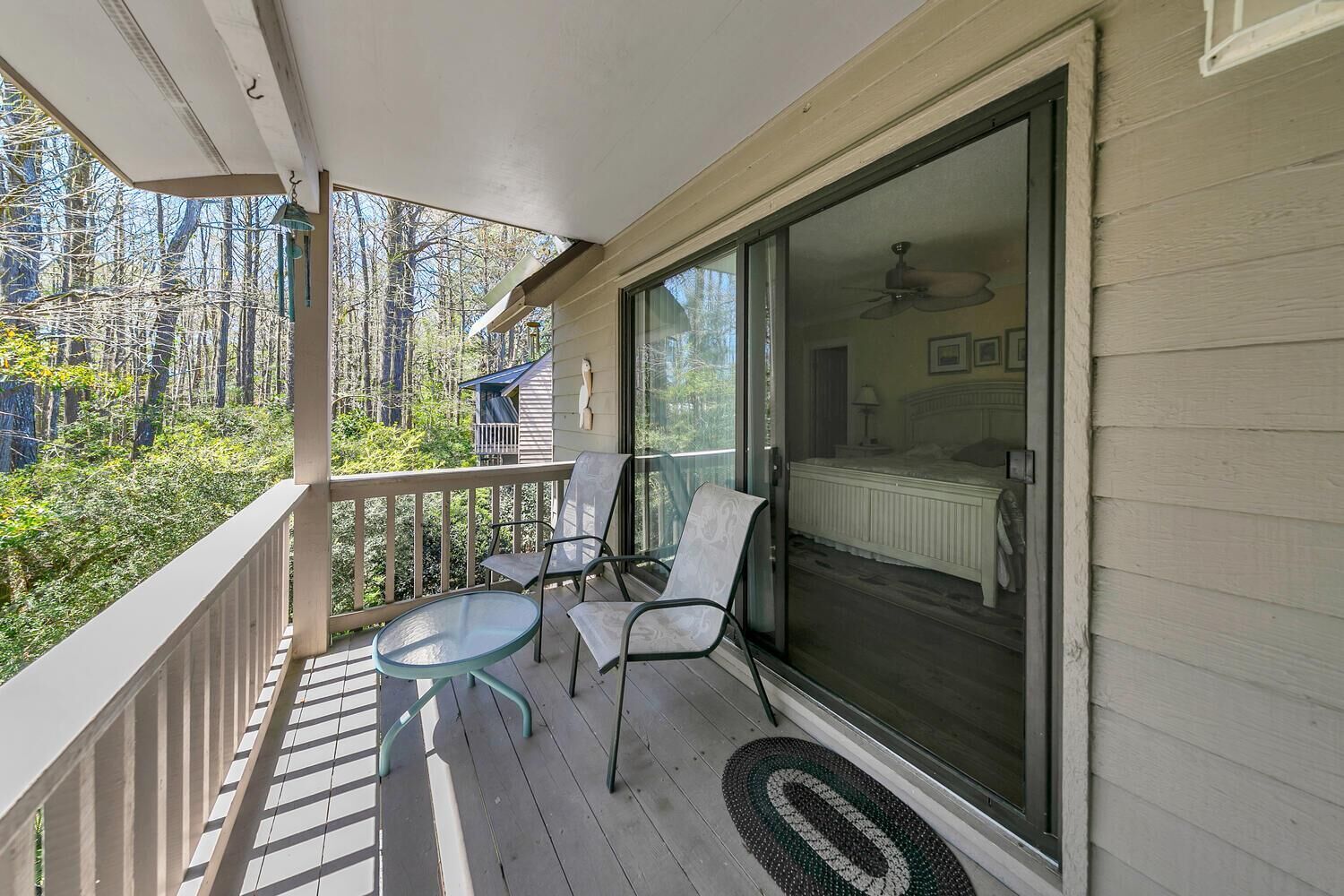 Un balcon avec une porte-fenêtre donnant sur une chambre, une petite table, deux chaises et une vue sur des arbres et un chalet.