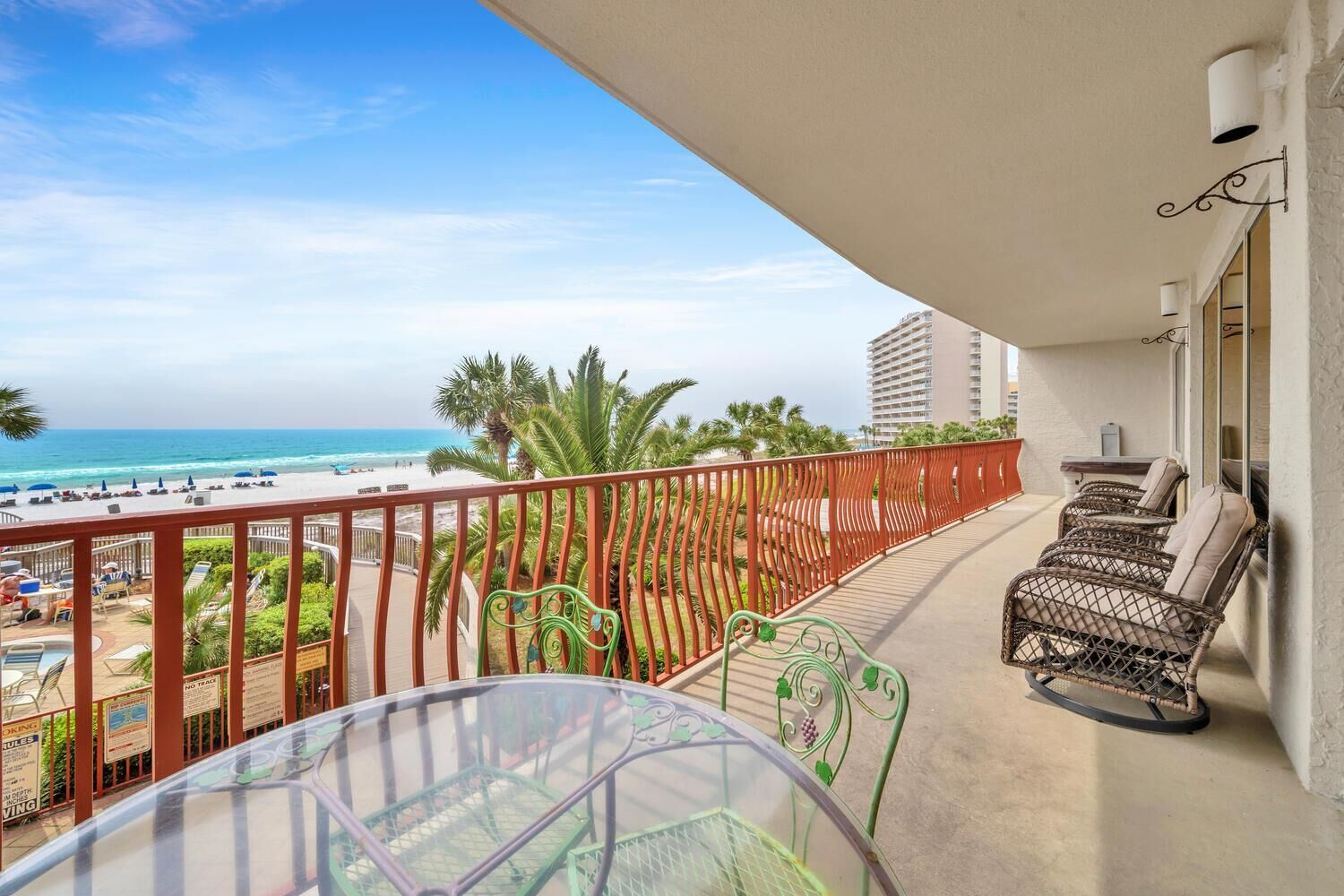A balcony with a glass table and chairs, overlooking a beach with palm trees and a hotel building in the background.