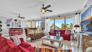 A spacious living room with a red sofa, a tufted armchair, and a red couch. There are ceiling fans, a coffee table, and a view of the outdoors through large windows. - Hidden Dunes 203 (Panama City Beach)