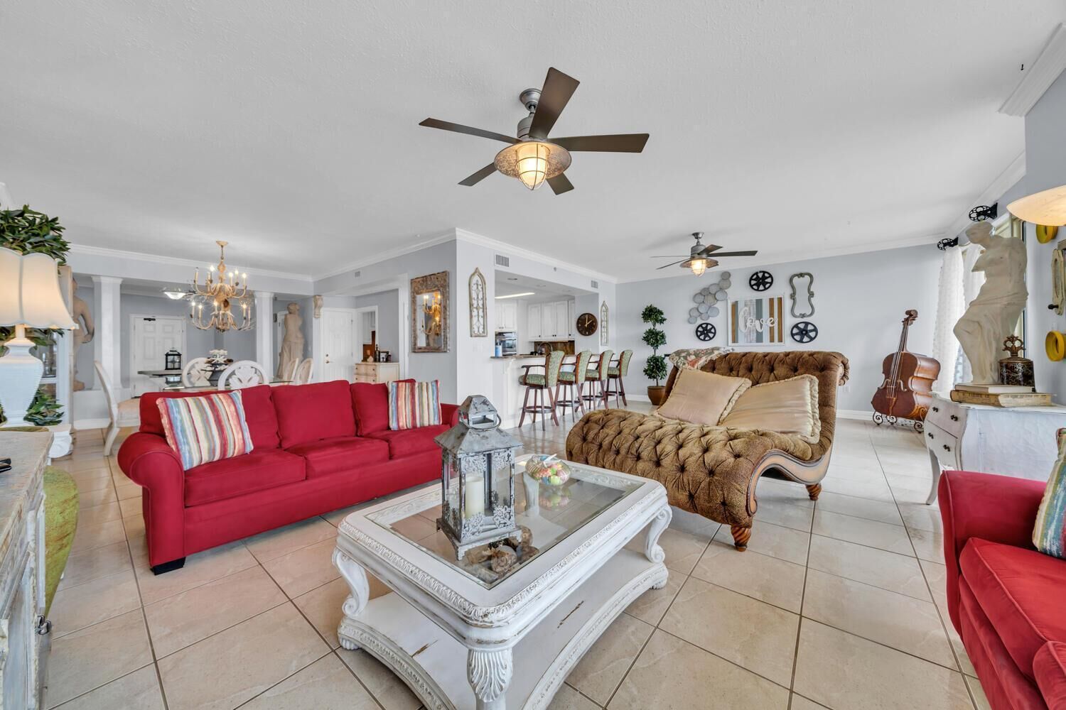Un salon spacieux avec un canapé rouge, un fauteuil marron et une table basse blanche. On y trouve deux ventilateurs de plafond, un lustre et un violon posé sur un pupitre.