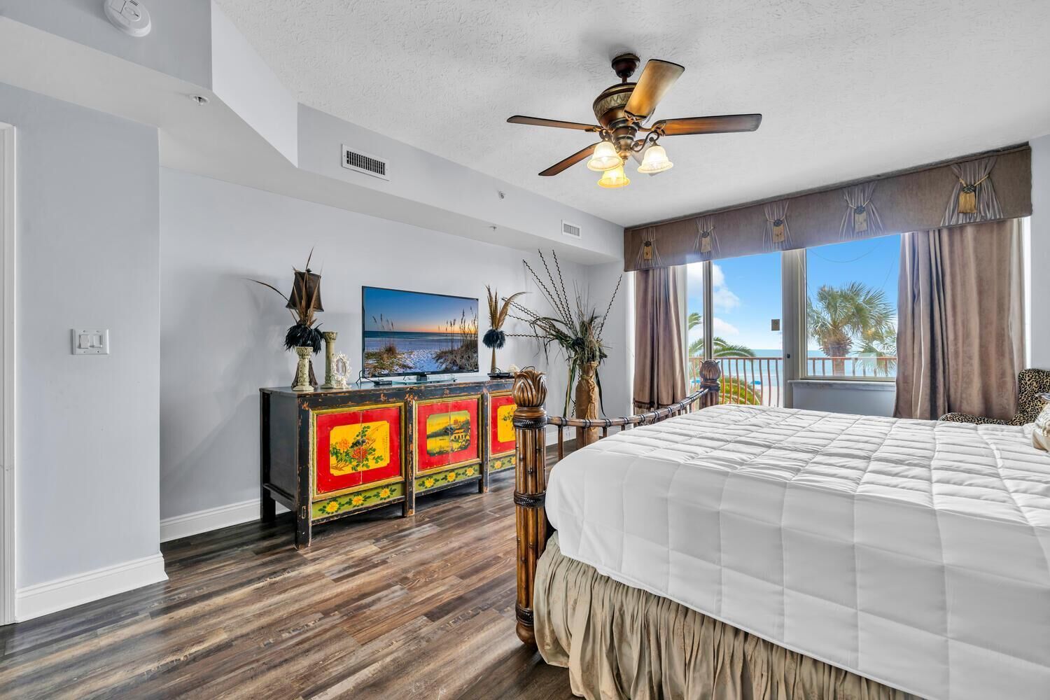A bedroom with a large bed, a ceiling fan, a TV, and a view of the beach.
