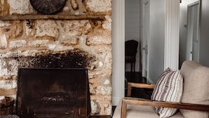 Une cheminée en pierre avec un foyer en métal, une chaise en bois et un canapé agrémenté de coussins à motifs, le tout dans une pièce dont un des murs est en pierre.