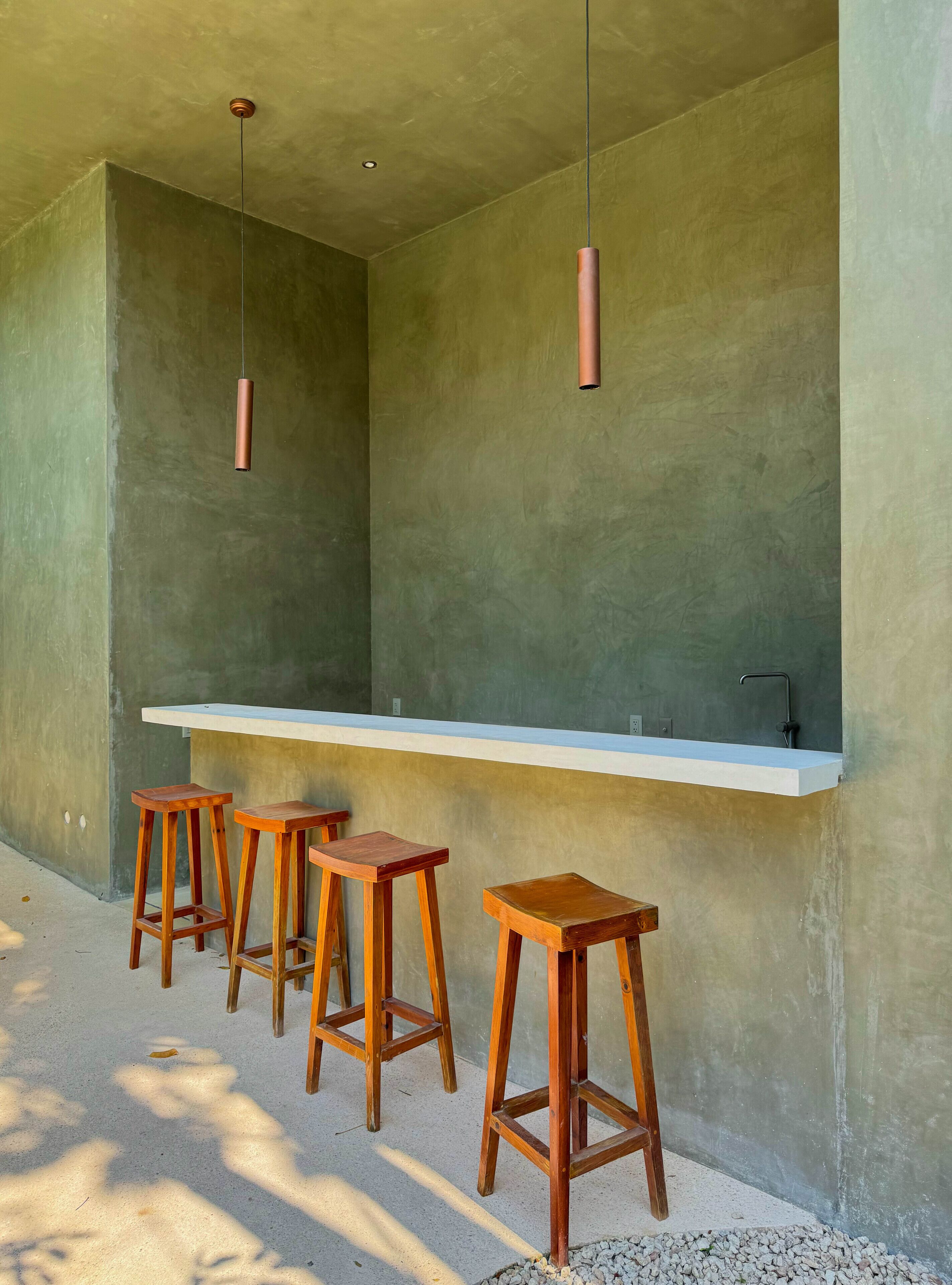 A bar area with a concrete wall, a white countertop, and three wooden stools.