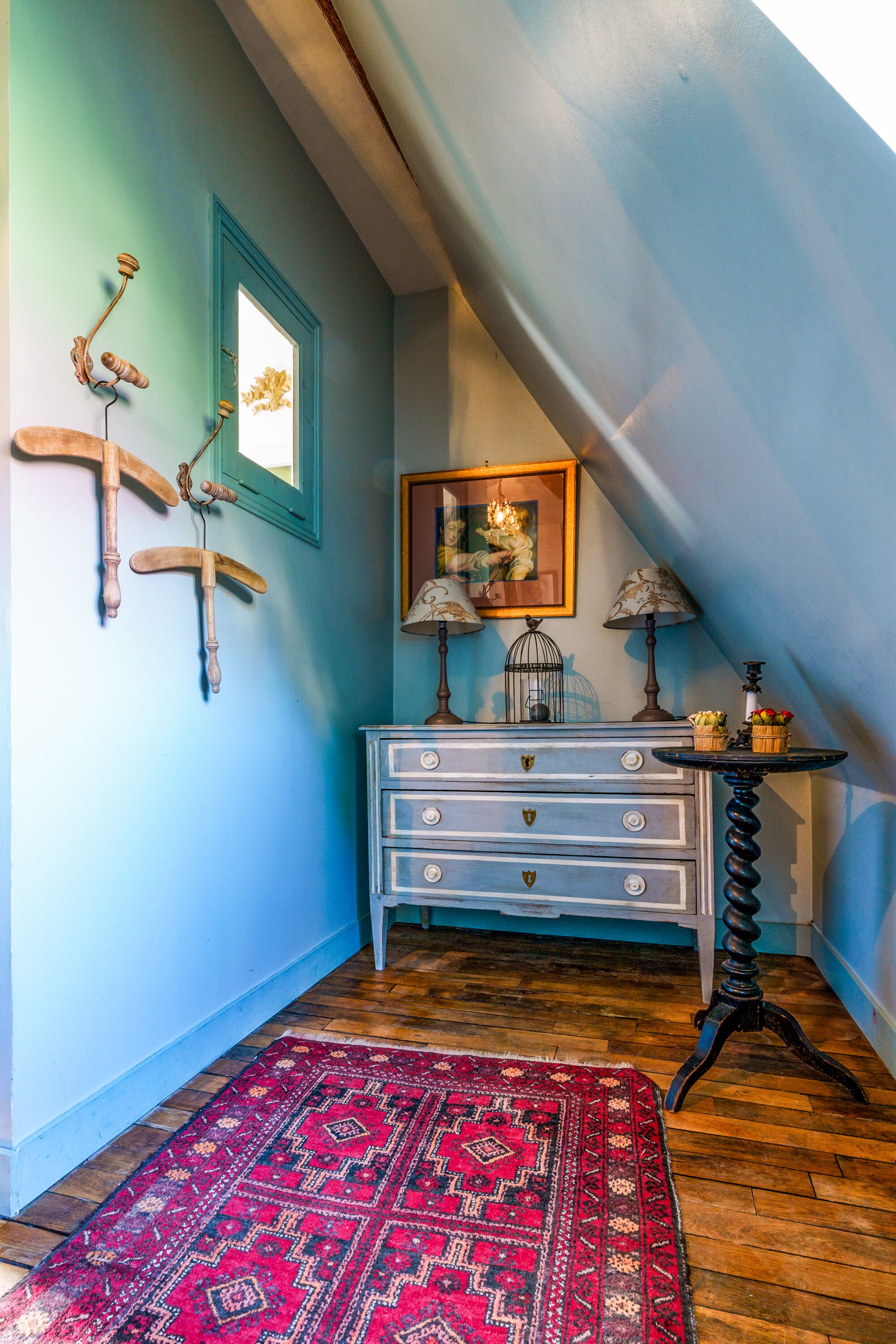 Une pièce avec un mur bleu, un plancher en bois, un tapis rouge à motifs, une commode blanche avec des tiroirs, une table noire avec une lampe et un tableau encadré accroché au mur.