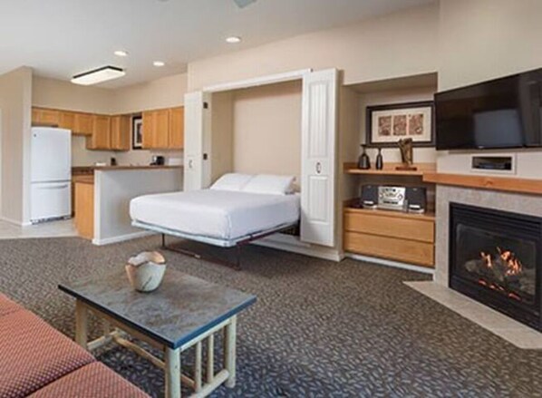 Een compacte hotelkamer met een bed, kitchenette, flatscreen-tv, open haard en een klein tafeltje.