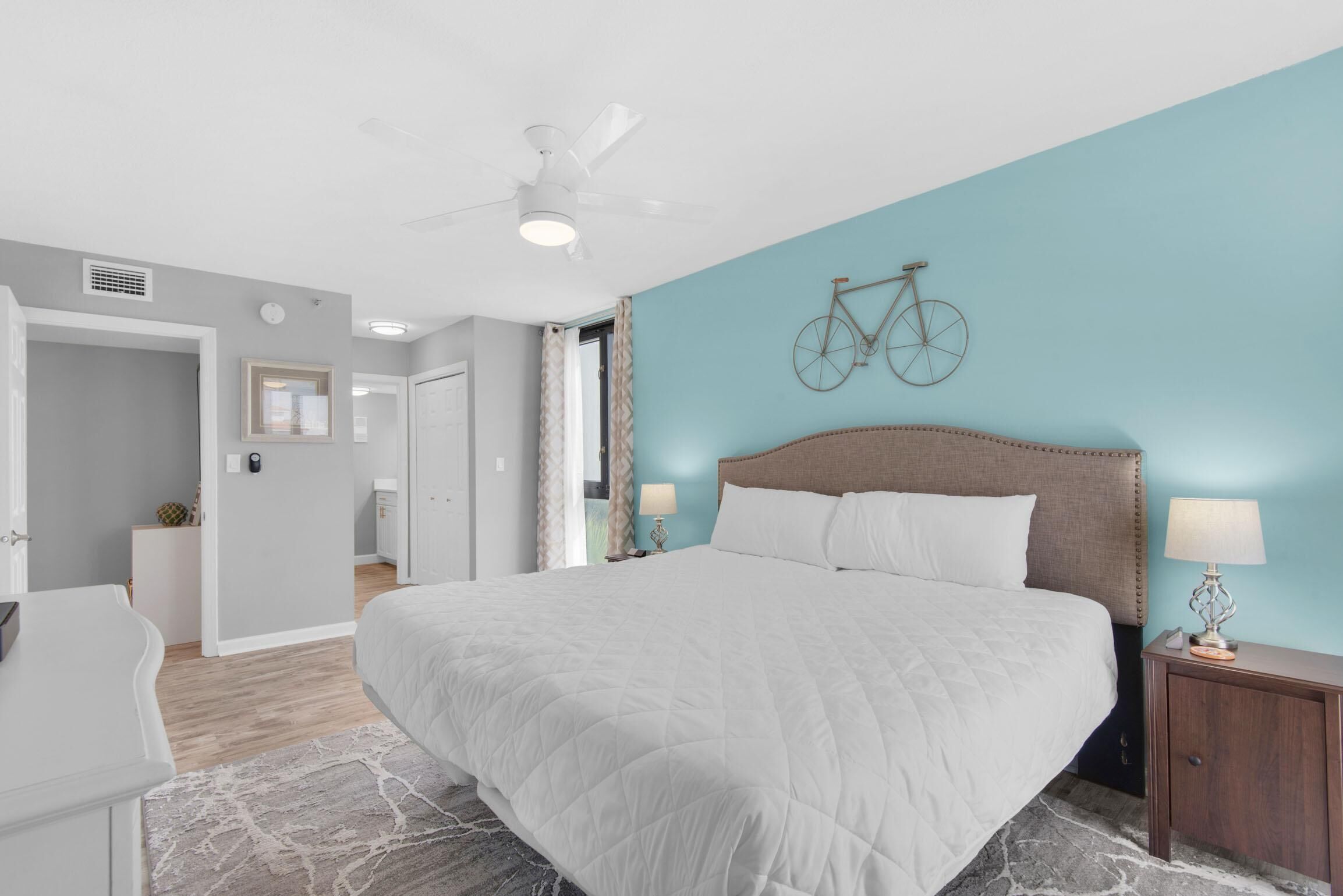 Une chambre avec un grand lit, un ventilateur de plafond, une décoration murale représentant un vélo et une table de chevet avec une lampe.