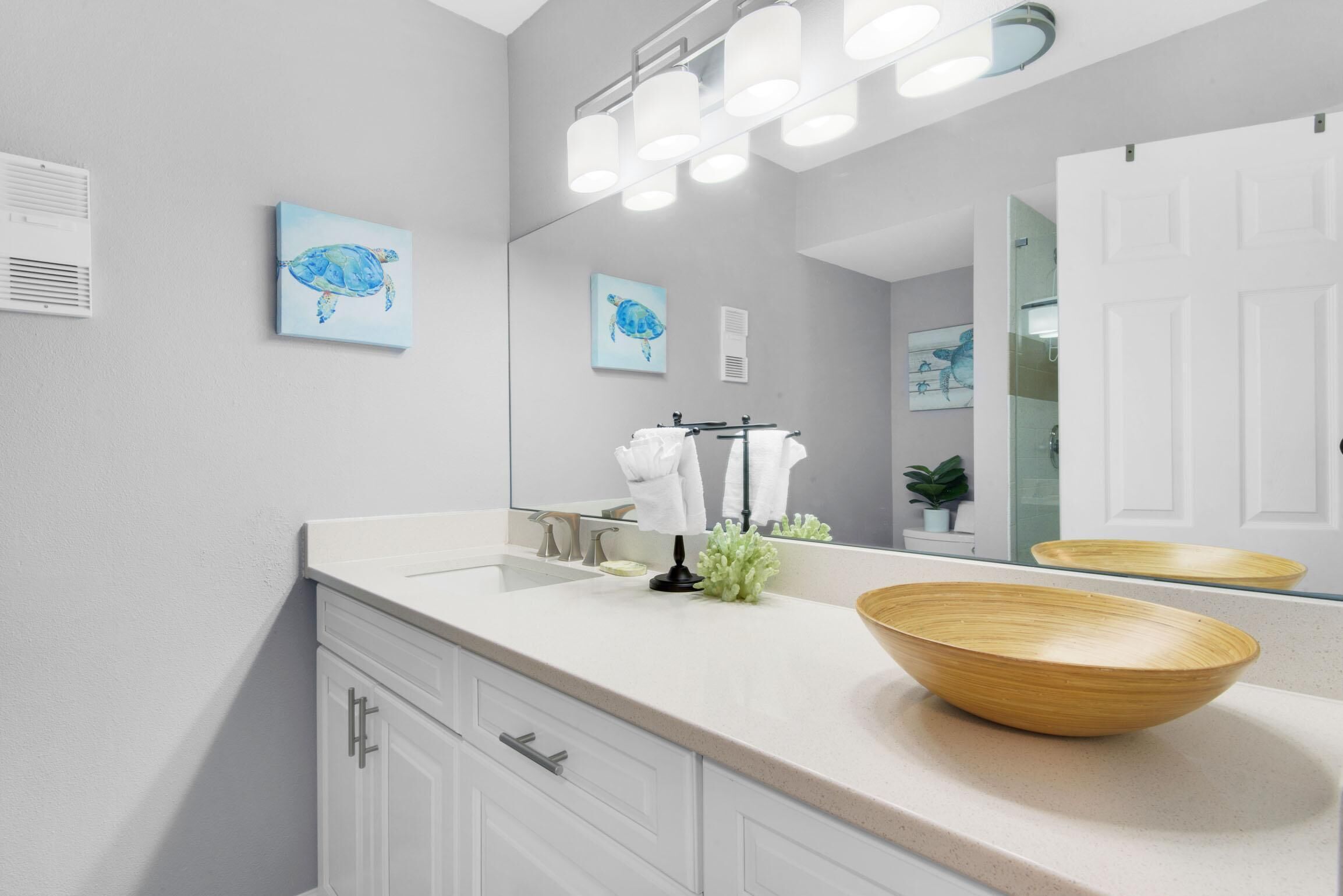 Une salle de bain moderne avec un grand miroir, deux œuvres d’art encadrées représentant des tortues de mer, un bol en bois posé sur le comptoir et une porte blanche.