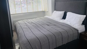 Un dormitorio con cama, ropa de cama blanca, un radiador y una ventana con persianas.