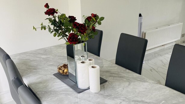 Una mesa de comedor con un florero de rosas rojas, dos rollos de papel higiénico y un cuenco de nueces.