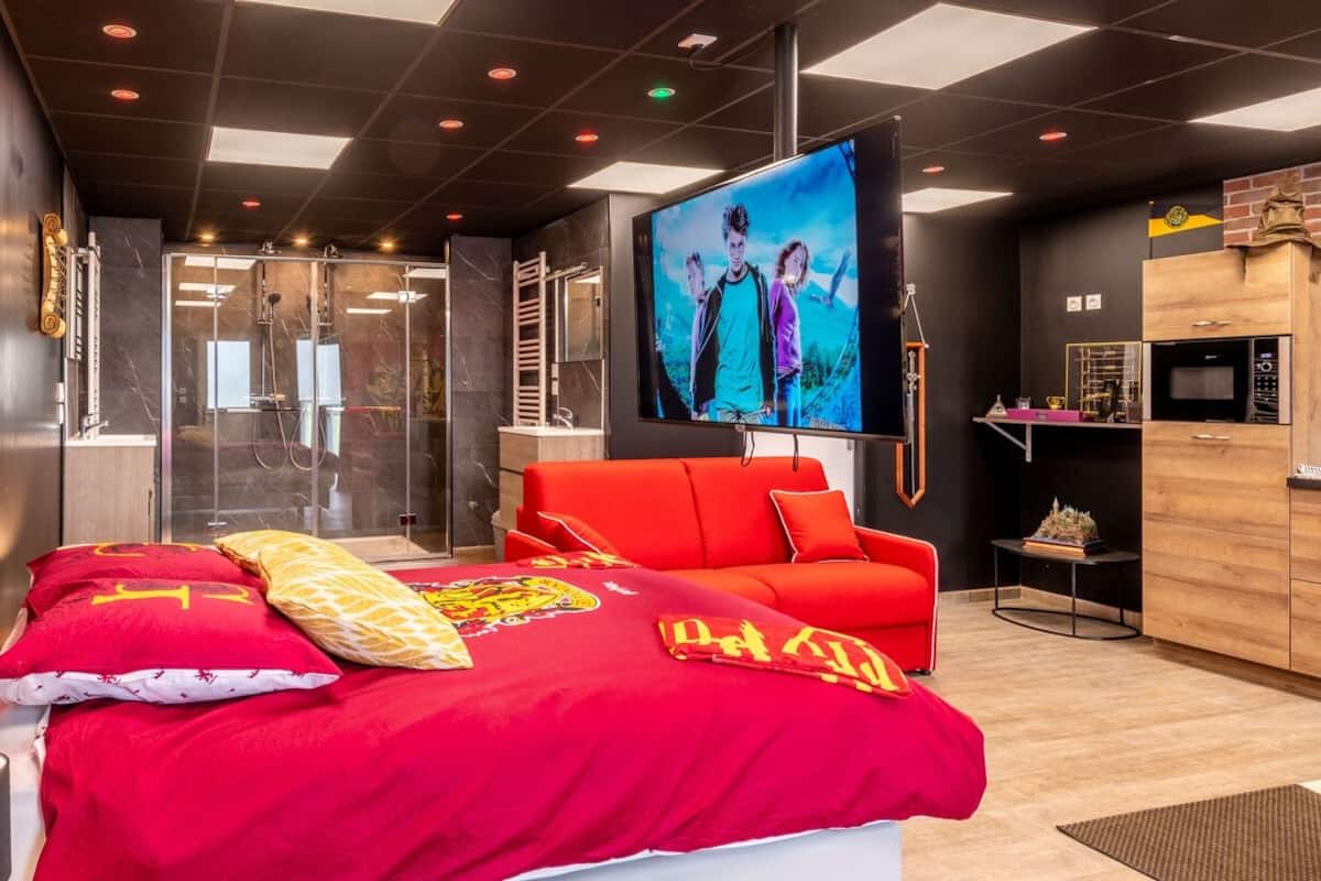 A modern hotel room with a large bed, a red sofa, a television, and a minibar.