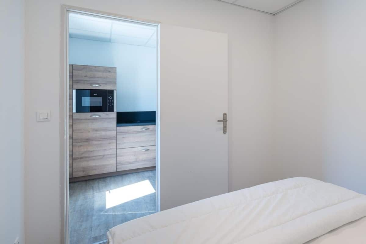 A bedroom with a bed, a wardrobe, and a kitchenette area visible through an open door.