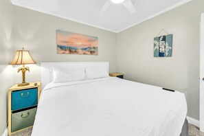 A bedroom with a bed, a nightstand with a lamp, a painting, and a wall decoration. - Misty Harbour 7 (Panama City Beach)