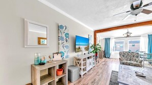 A spacious living area with a wooden floor, a large window with curtains, a ceiling fan, and a view of the beach. - Misty Harbour 7 (Panama City Beach)
