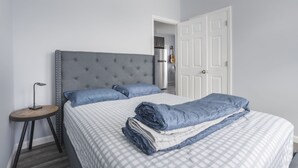 Une chambre à coucher avec une tête de lit capitonnée grise, un lit recouvert d’une couette à carreaux, une petite table ronde avec une lampe, et une porte blanche en arrière-plan.