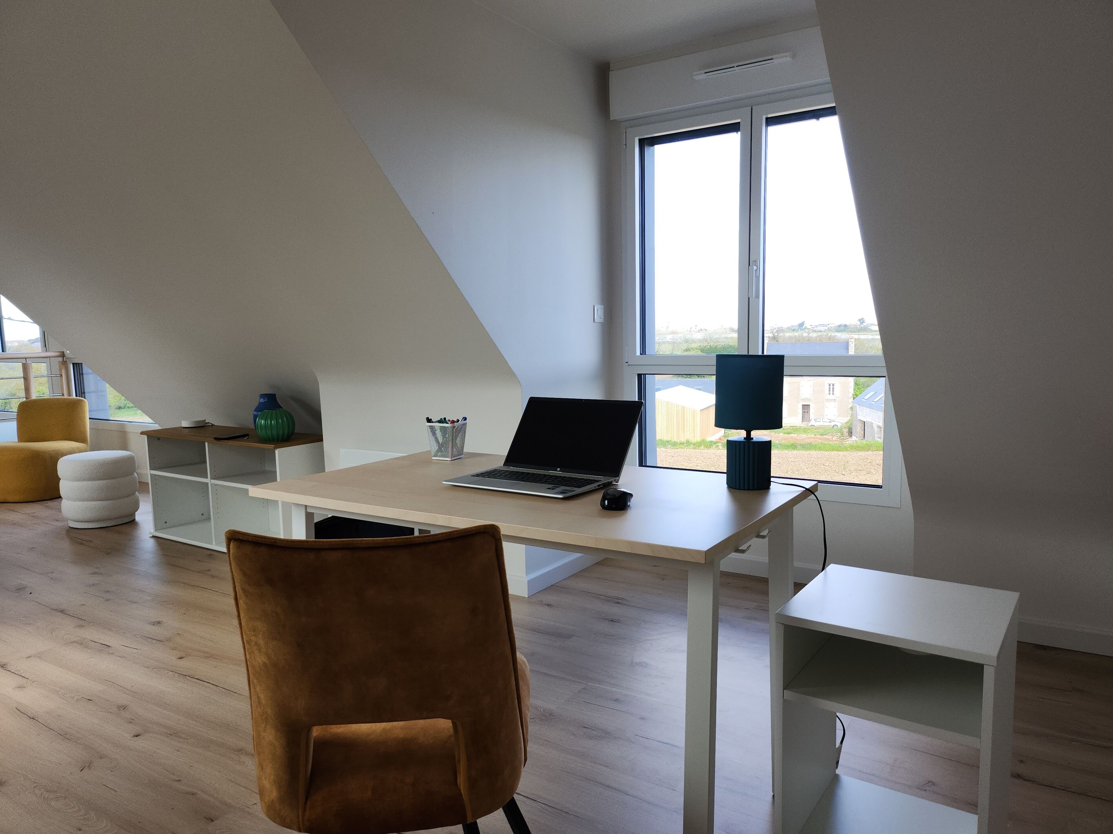 A modern, well-lit room with a sloped ceiling, a desk with a laptop, a chair, and a view of buildings outside through large windows.