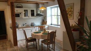 A kitchen with wooden beams, white cabinets, a dining table with chairs, and a window with a sunflower painting. - Ferienwohnung Direkt am See (Wokuhl-Dabelow)