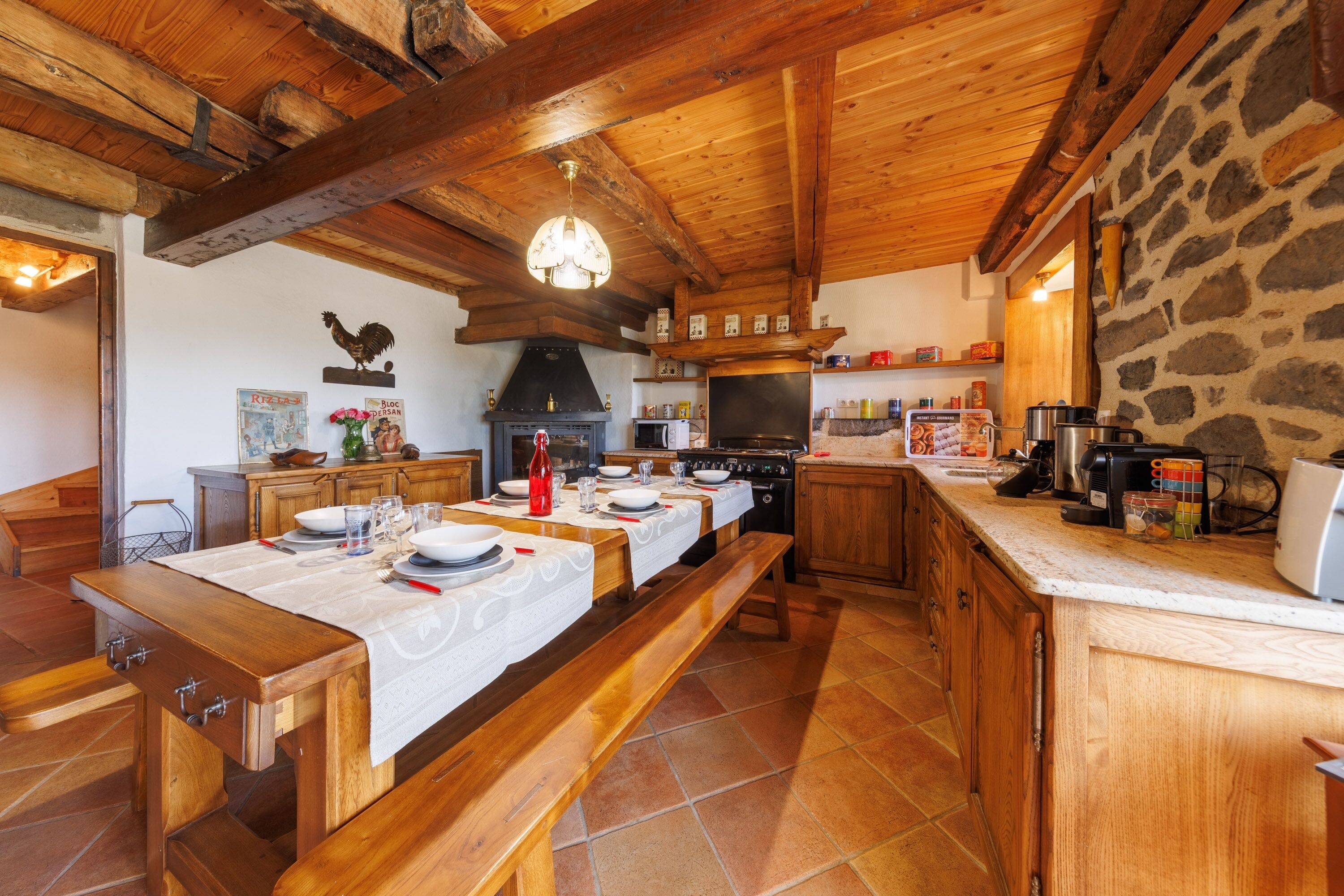 A rustic kitchen with wooden beams, a stone wall, and a long dining table set for four.