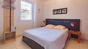 Un dormitorio con una cama, una mesita de noche con lámpara, un estante de madera, un ventanal y una pared decorada con marcos.
