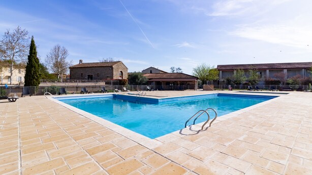 Una piscina rectangular con solárium alicatado, rodeada por una valla, y contigua a edificios con tejados de teja.