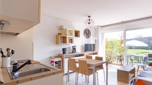 Cocina moderna con lavabo, mesa de comedor, sillas y televisor. Se ve un ventanal con vistas a zonas verdes.