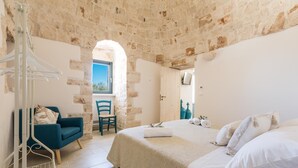 A bedroom with a stone wall, a bed with white bedding, a blue chair, and a white hanging rack.