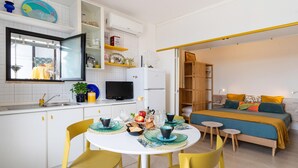 Cocina compacta con armarios blancos, una mesa de comedor con sillas amarillas y una cama con colcha azul y naranja en la habitación contigua.