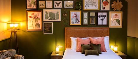 Un dormitorio con una cama grande, varias láminas botánicas enmarcadas en la pared, una mesita de noche con una lámpara y un sillón con estampado tropical.