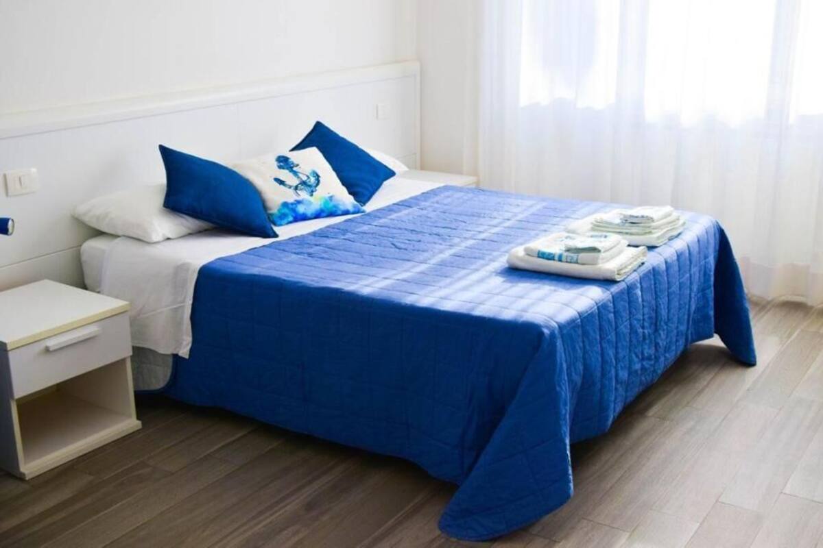 A neatly made bed with a blue bedspread and pillows, a white bedside table, and a window with sheer curtains.